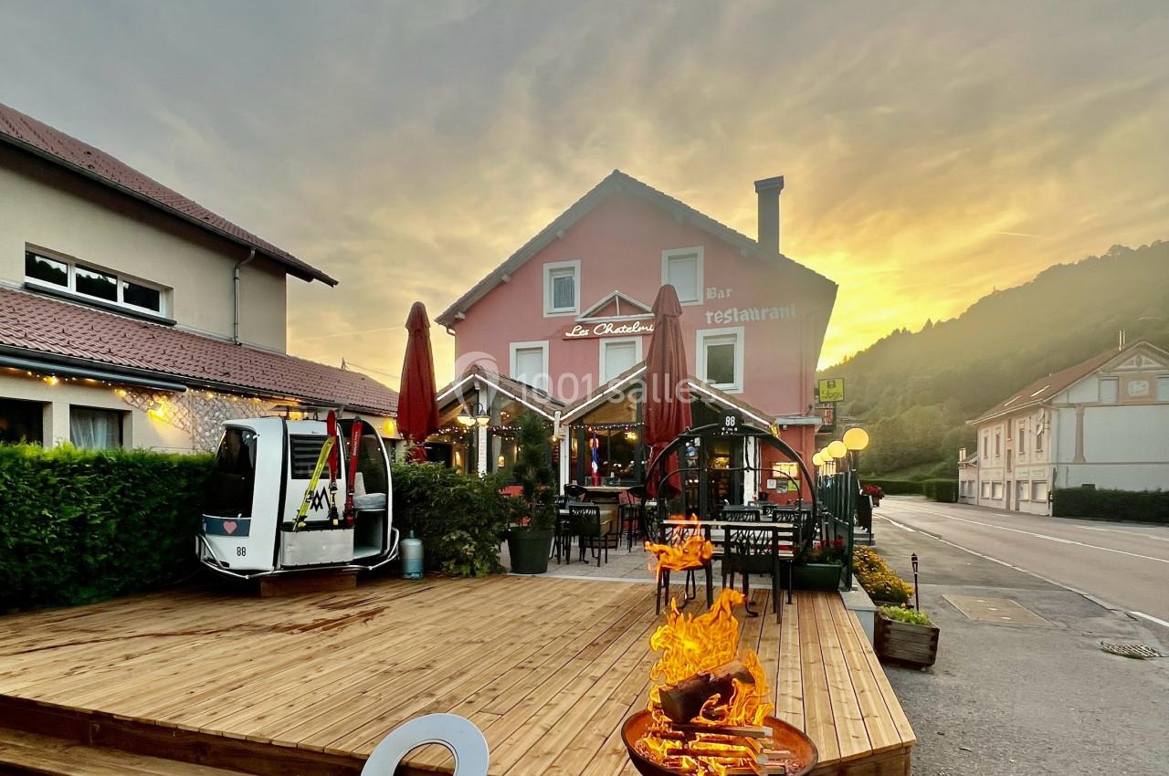 Terrasse en bois d'un restaurant avec brasero allumé, parasols rouges et bâtiment rose au coucher du soleil.