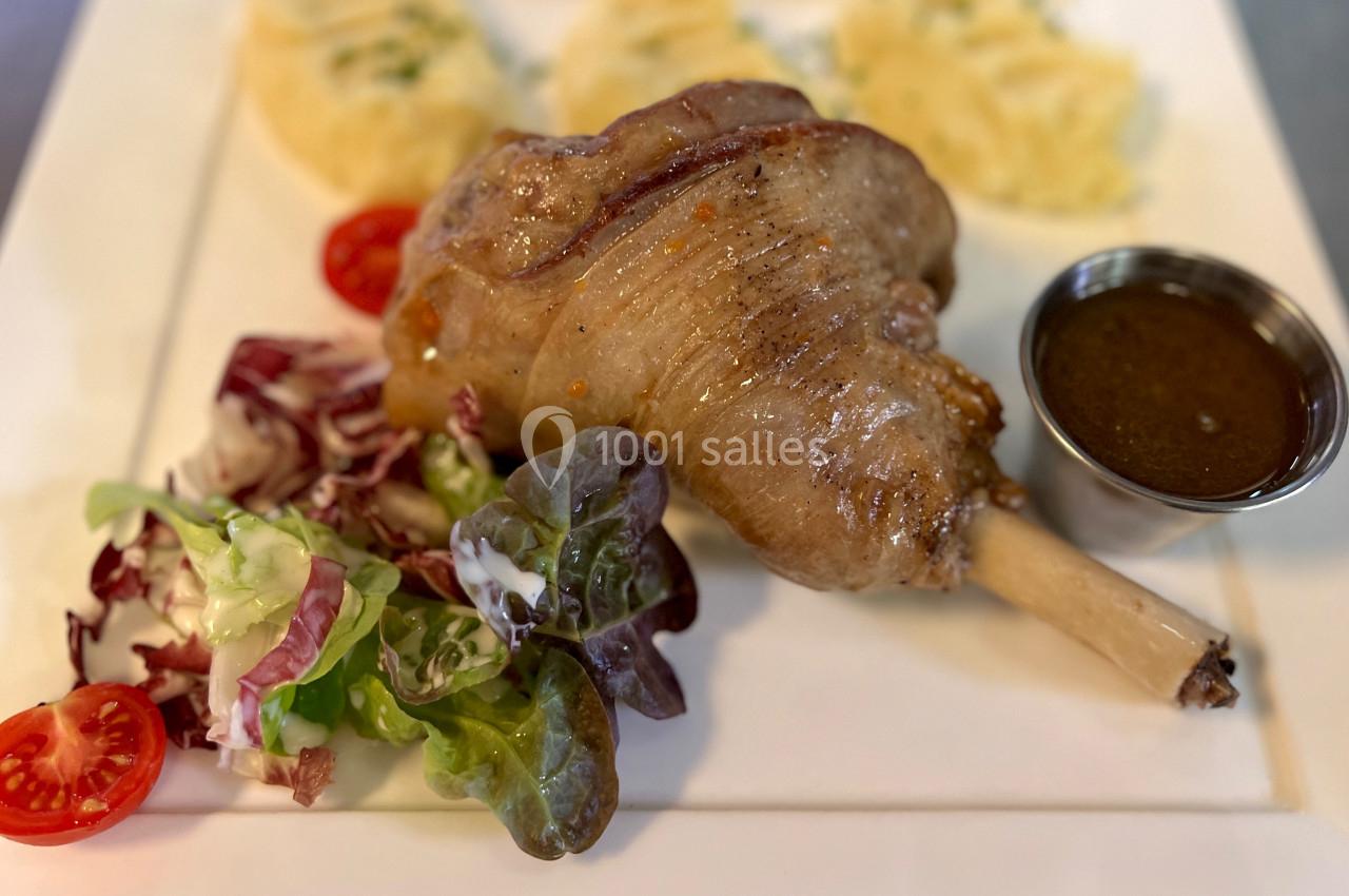 Jarret d'agneau rôti servi avec purée de pommes de terre, salade verte et sauce dans une coupelle en métal.