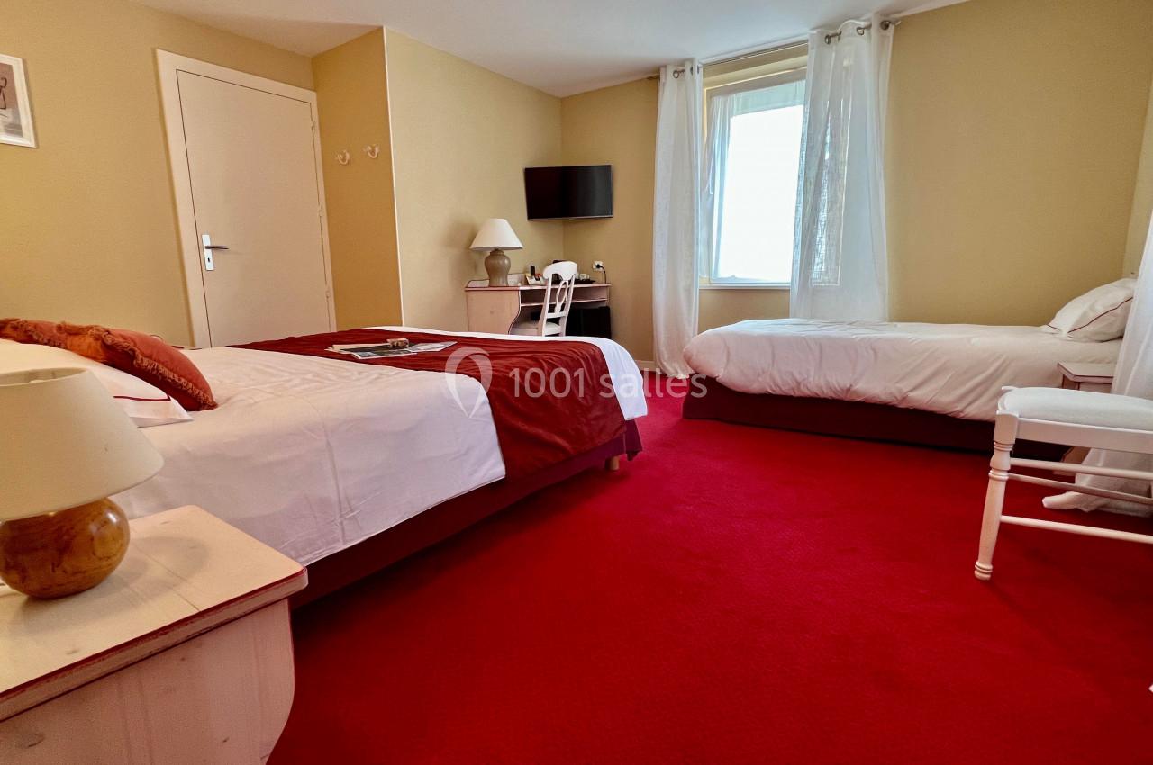 Chambre avec deux lits, moquette rouge, bureau avec chaise, télévision murale et fenêtre laissant entrer la lumière.