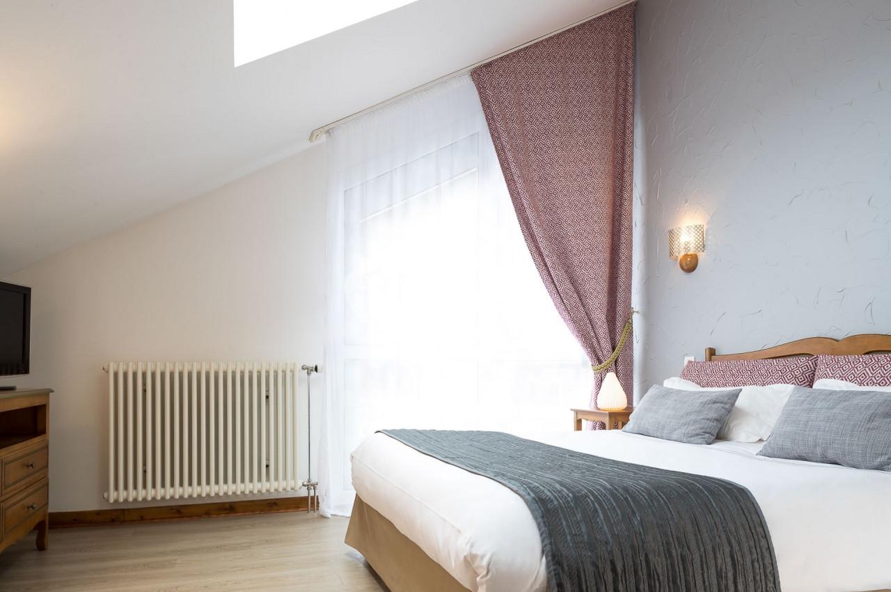Chambre lumineuse avec lit double, rideaux rouges, mur gris, radiateur blanc et mobilier en bois clair.