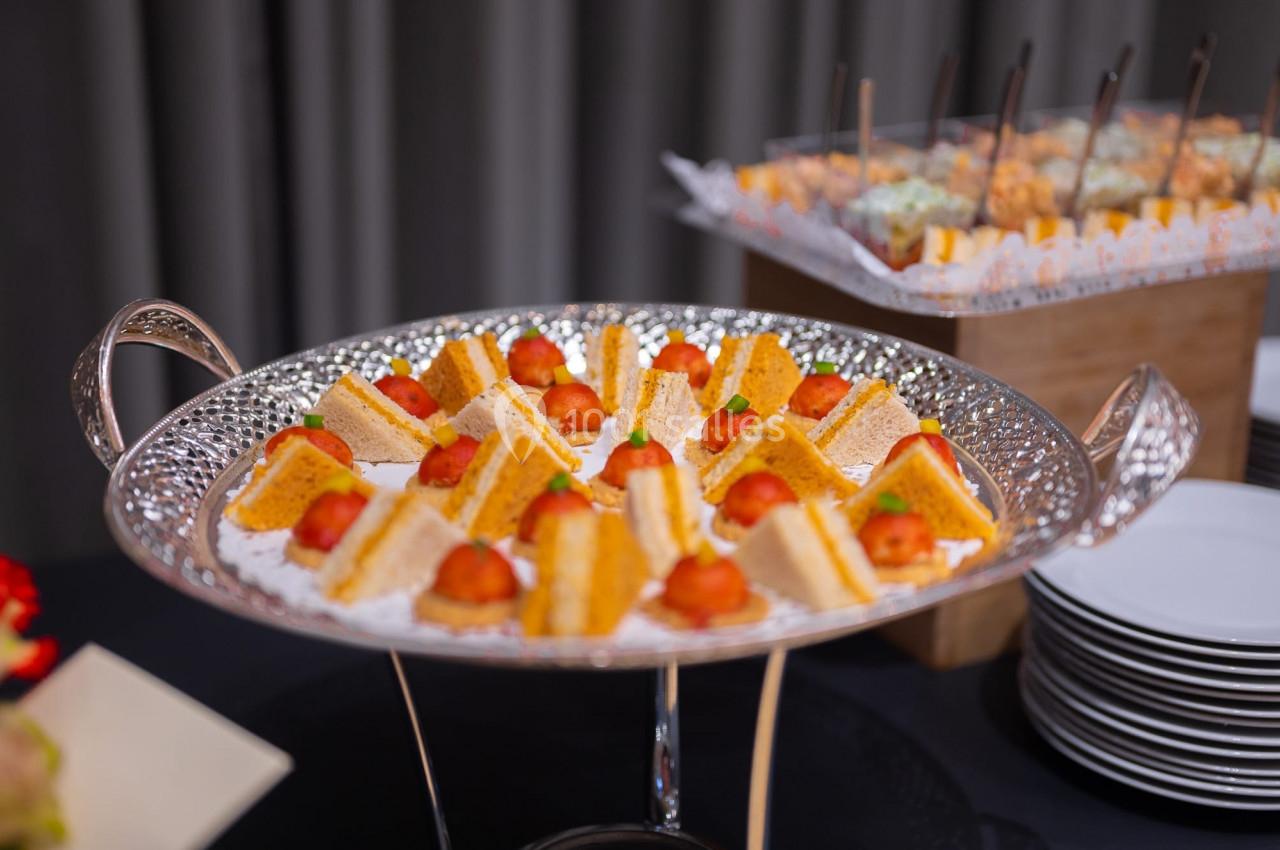 Plateau argenté contenant des amuse-bouches triangulaires garnis de tomates cerises, avec d'autres plats en arrière-plan.