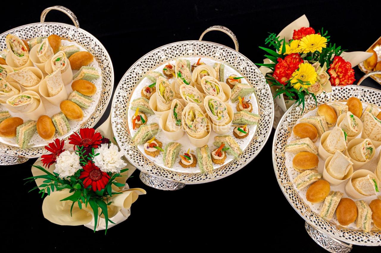 Plateaux garnis de sandwichs roulés, mini-sandwichs et amuse-bouches, entourés de bouquets de fleurs colorées.