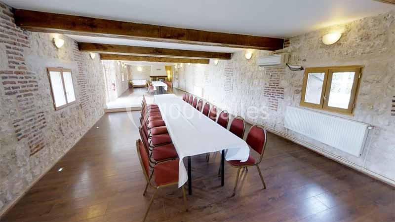 Salle lumineuse avec murs en pierre, poutres apparentes, table rectangulaire dressée et chaises rouges.