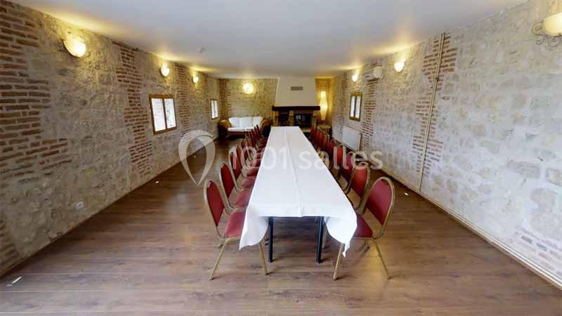 Salle en pierre avec un long table rectangulaire dressée, entourée de chaises rouges, éclairée par des appliques murales.
