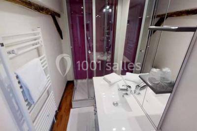 Salle de bain moderne avec douche vitrée, lavabo blanc, miroir, sèche-serviettes et poutres en bois apparentes.