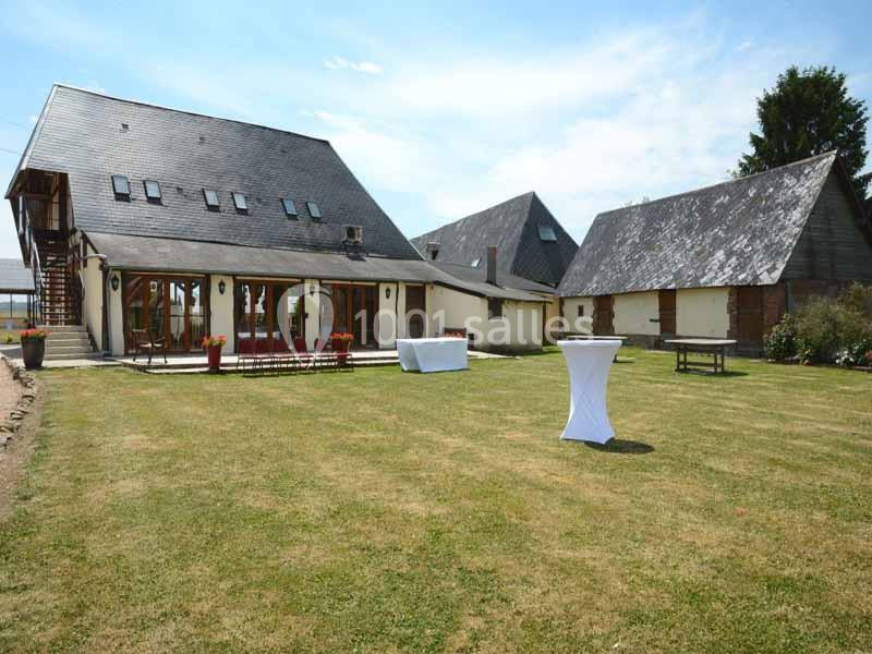 Grande maison avec toit en ardoise entourée de pelouse, tables dressées et bâtiments annexes en arrière-plan.