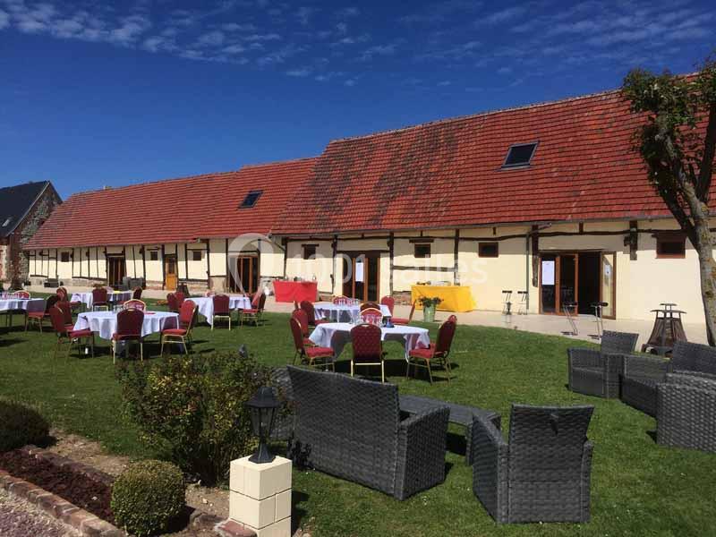 Tables dressées et fauteuils en osier disposés sur une pelouse devant un bâtiment à toit rouge par temps ensoleillé.