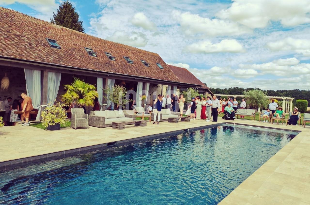 Groupe de personnes rassemblées près d'une piscine dans un jardin, devant un bâtiment en pierre avec toit en tuiles.
