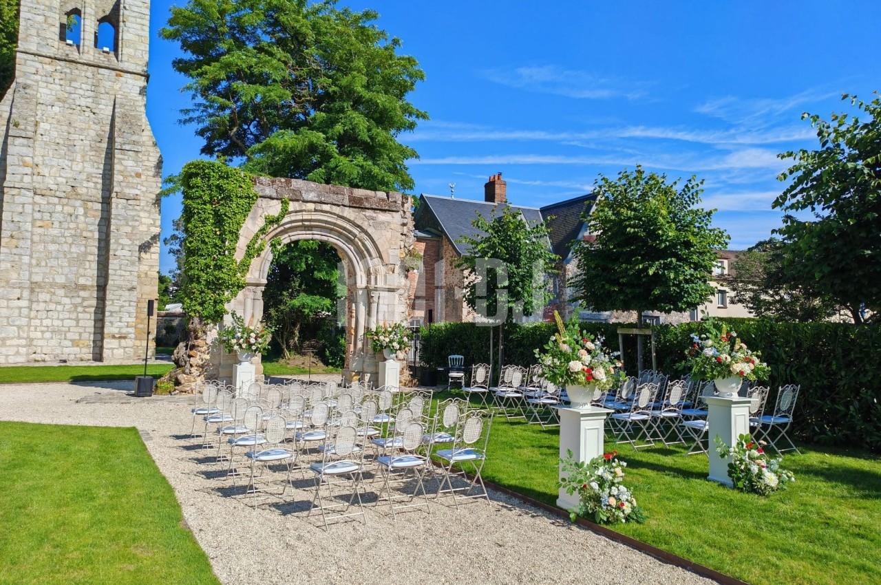 Chaises alignées devant une arche en pierre dans un jardin, préparées pour une cérémonie en plein air par temps ensoleillé.