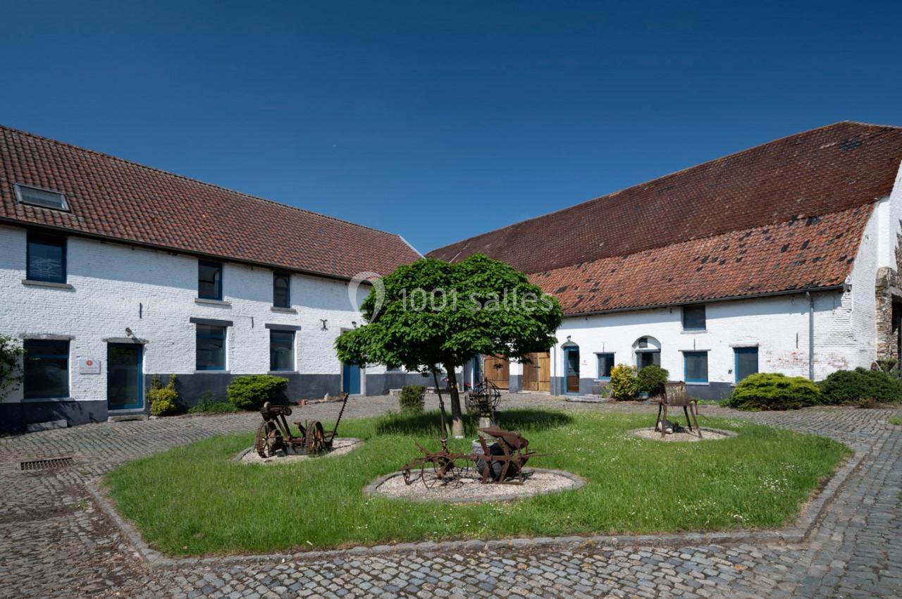 Cour intérieure pavée avec pelouse centrale, arbre, anciens outils agricoles et bâtiments en briques blanches.