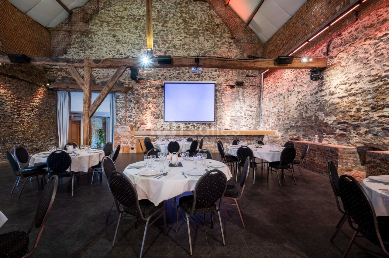 Salle de réception avec murs en pierre, tables rondes dressées et un écran de projection au fond.