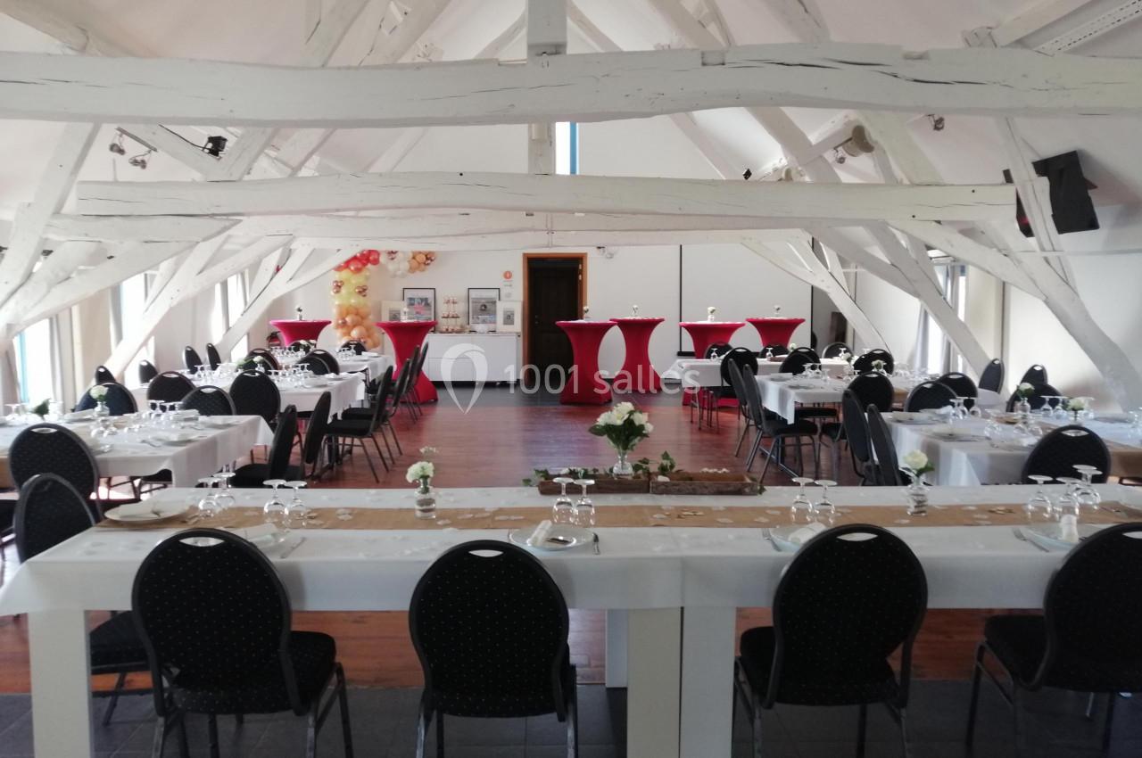 Salle lumineuse avec poutres apparentes, tables dressées pour un événement, chaises noires et décorations rouges.