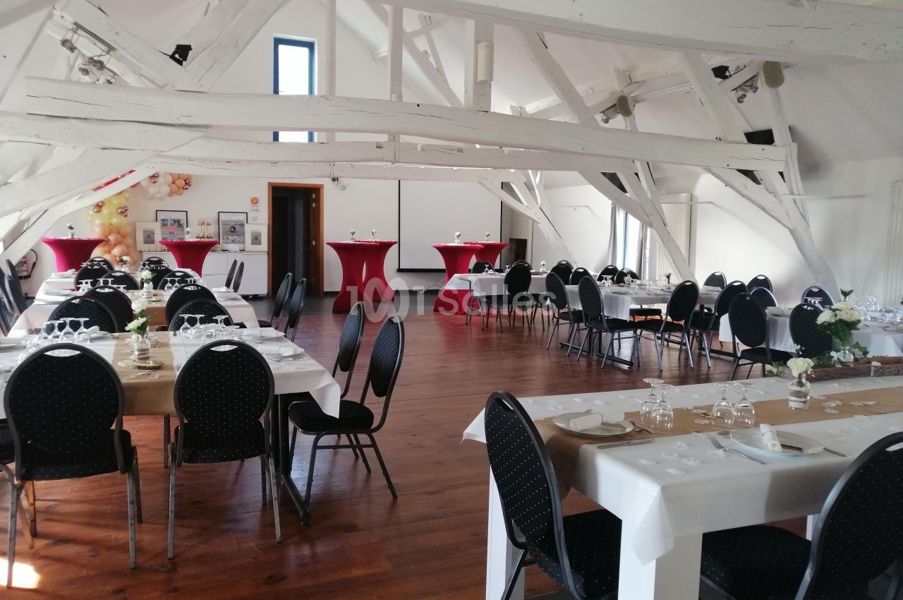 Salle lumineuse avec poutres apparentes, tables dressées pour un événement et chaises noires disposées autour.