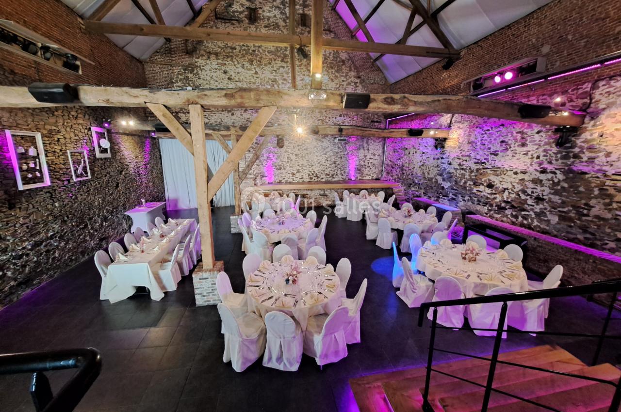 Salle de réception en pierre avec poutres apparentes, tables rondes dressées et éclairage violet.