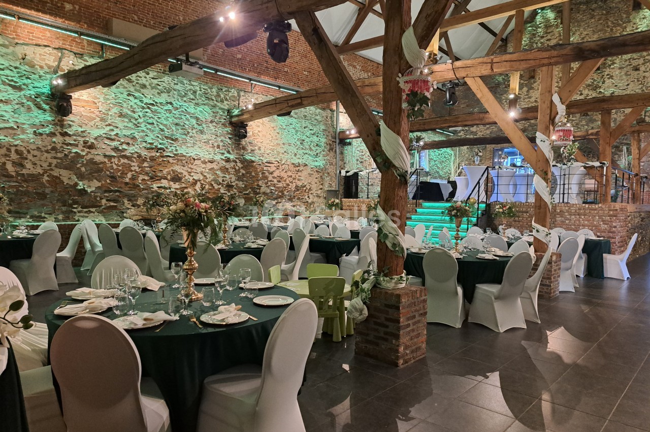 Salle de réception décorée avec des tables rondes dressées, chaises blanches et éclairage vert sur des murs en pierre.