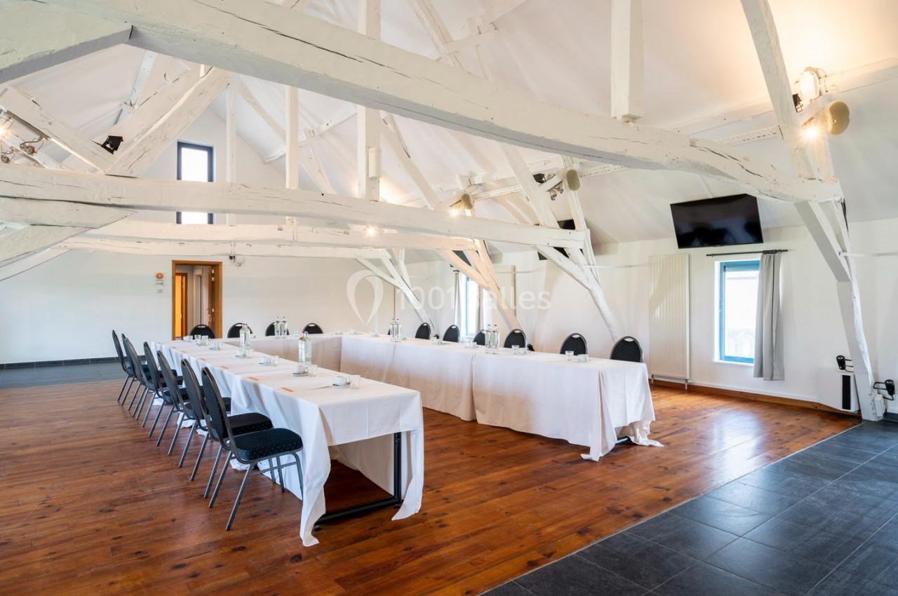 Salle de réunion lumineuse avec poutres apparentes, tables disposées en U, chaises noires et écran mural.