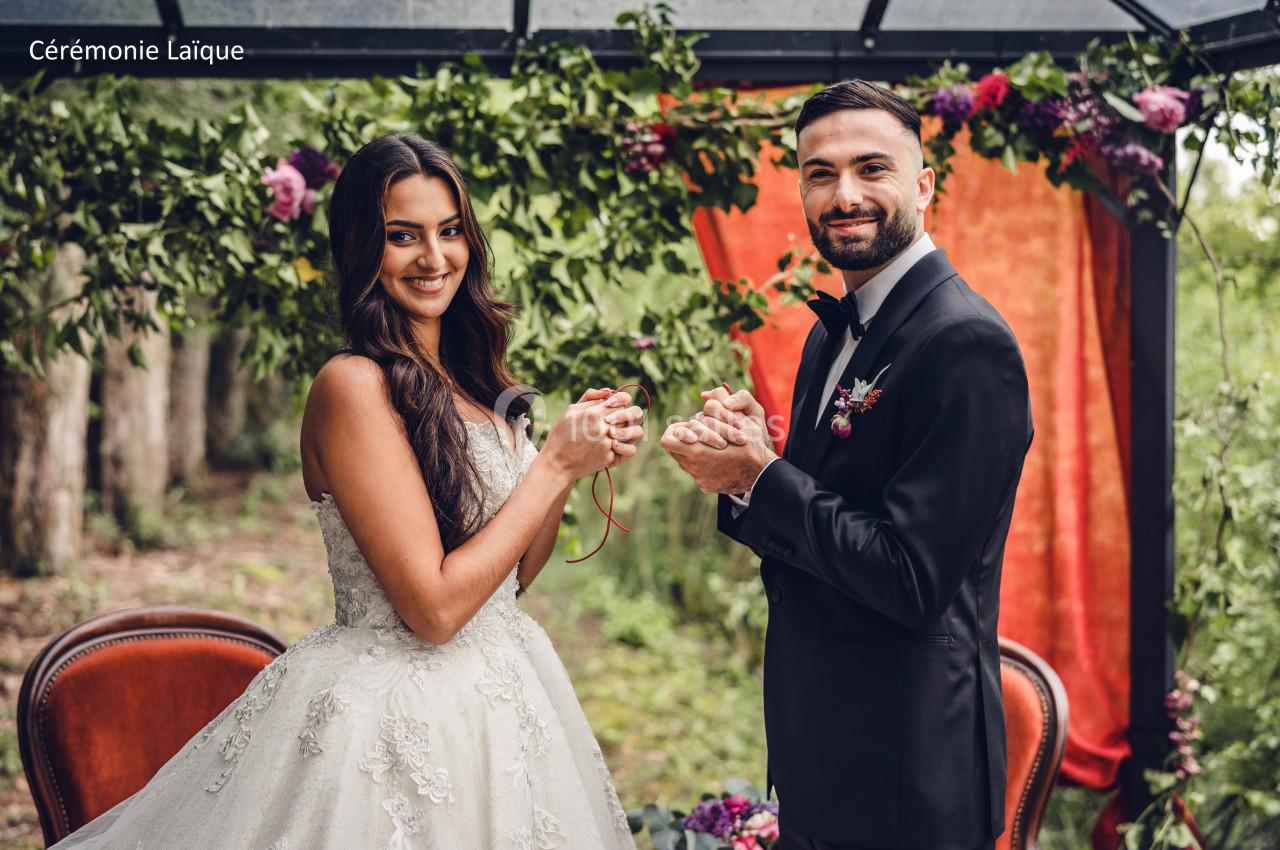 Un couple en tenue de mariage se tient les mains sous une arche décorée de feuillage et de fleurs.