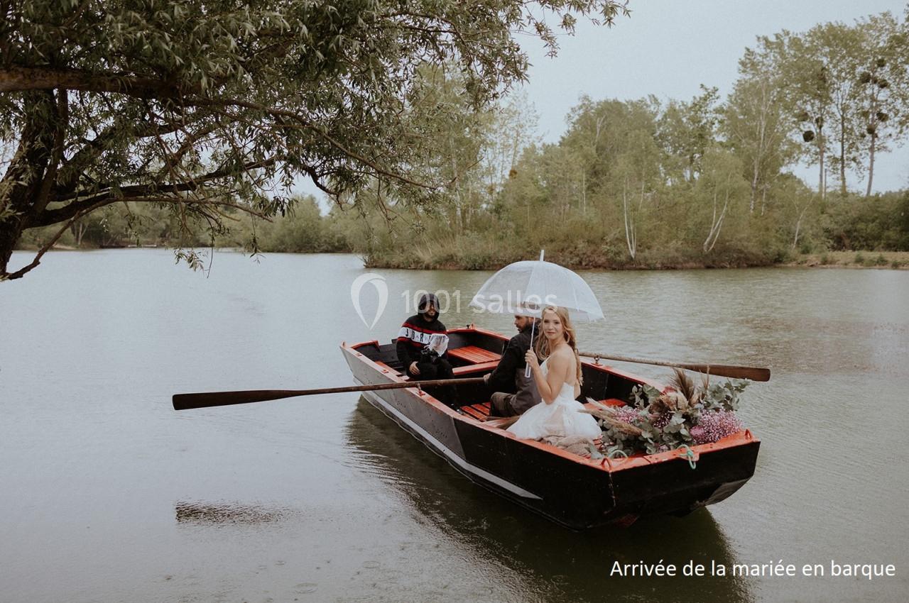 Une mariée en robe blanche arrive en barque sur un lac, accompagnée d'une personne ramant sous un ciel nuageux.