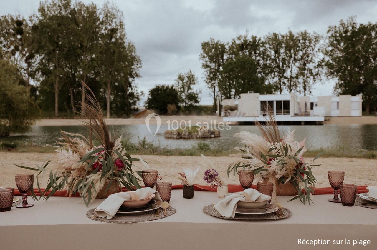 Table décorée avec des fleurs séchées et vaisselle élégante, installée en extérieur près d'un plan d'eau.