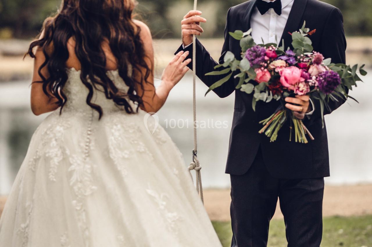 Un marié en costume tient un bouquet de fleurs face à une mariée en robe blanche près d'une balançoire en extérieur.