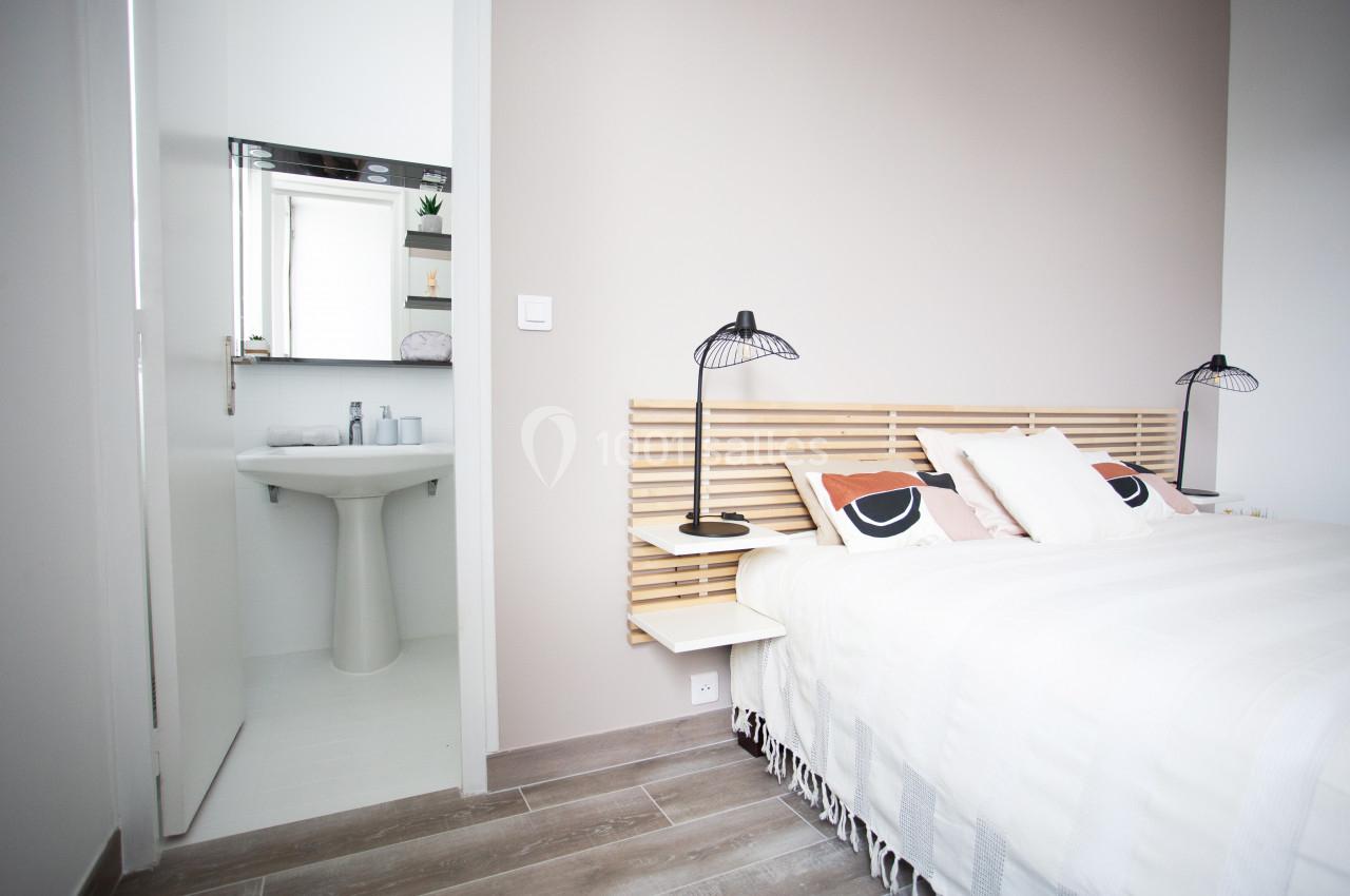 Chambre lumineuse avec lit double, tête de lit en bois clair, lampes noires et salle de bain attenante visible.