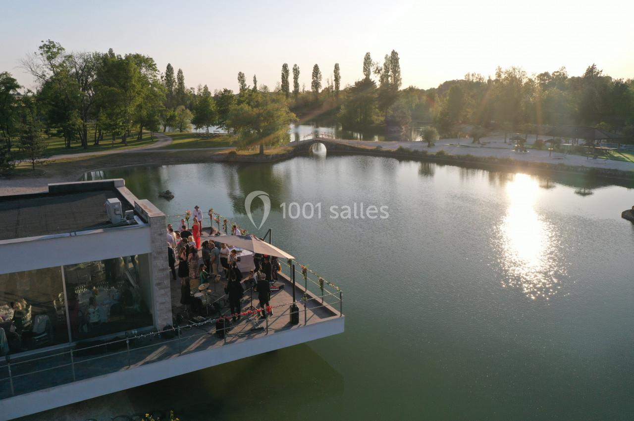 Vue aérienne d'un bâtiment avec terrasse surplombant un lac entouré d'arbres au coucher du soleil.
