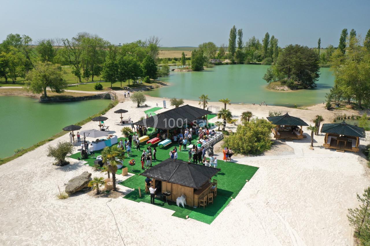 Vue aérienne d'une plage aménagée avec des kiosques, des parasols et des visiteurs près d'un lac entouré de verdure.