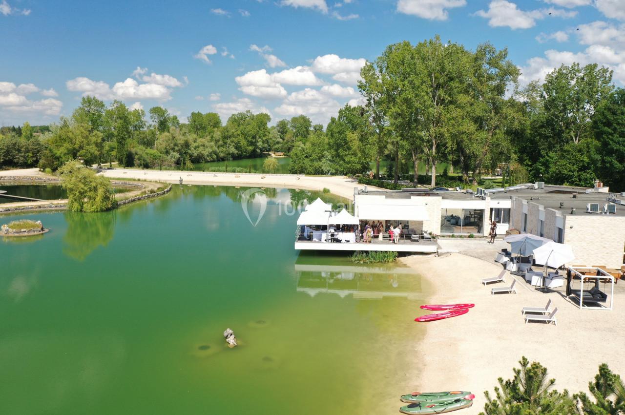 Vue aérienne d'un lac avec une plage, des kayaks sur le rivage, une terrasse aménagée et des arbres en arrière-plan.