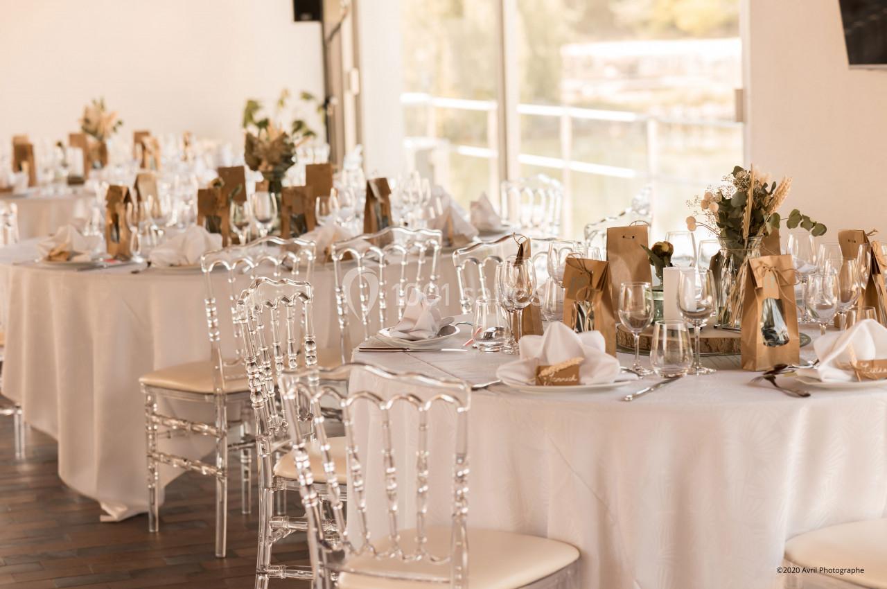 Tables élégamment dressées avec nappes blanches, chaises transparentes et décorations florales dans une salle lumineuse.