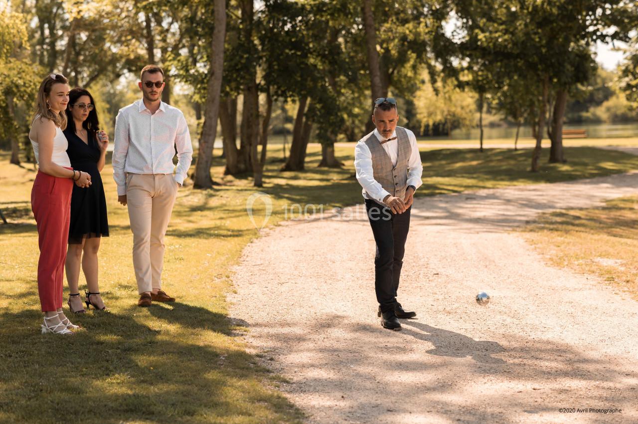 Un homme joue à la pétanque sur un chemin en gravier, observé par quatre personnes debout à l'ombre des arbres.