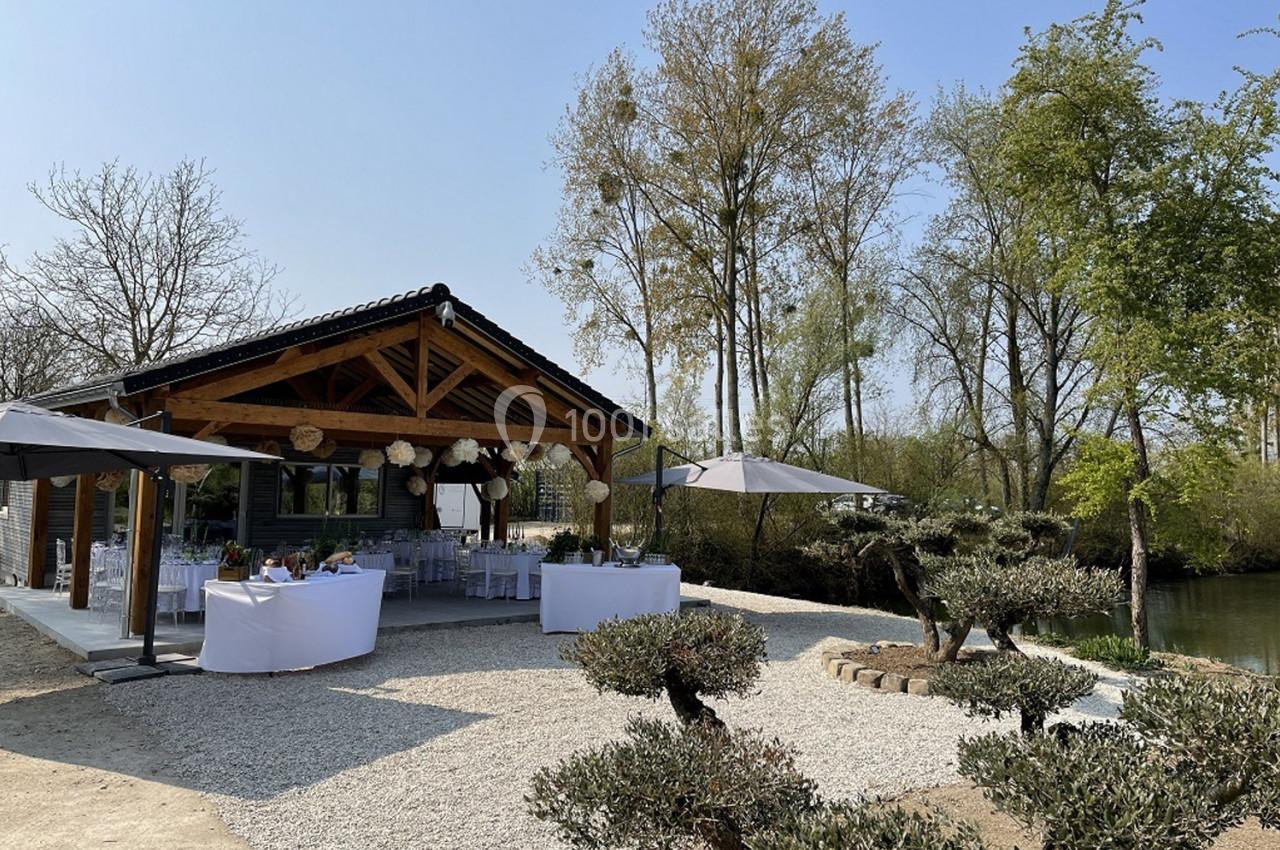 Pavillon en bois avec tables dressées pour un événement, entouré d'arbres et situé près d'un plan d'eau.
