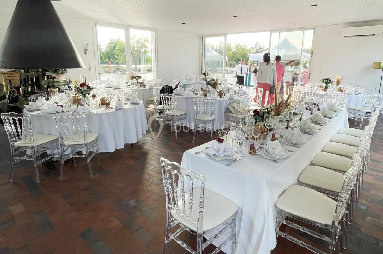 Salle de réception lumineuse avec tables dressées, chaises transparentes et vue sur l'extérieur.