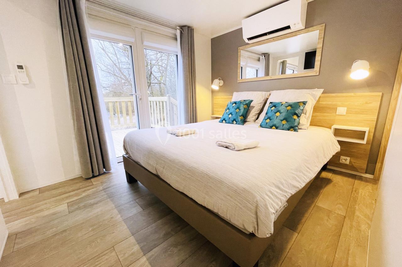 Chambre lumineuse avec lit double, coussins décoratifs, draps pliés, grande baie vitrée et sol en bois clair.