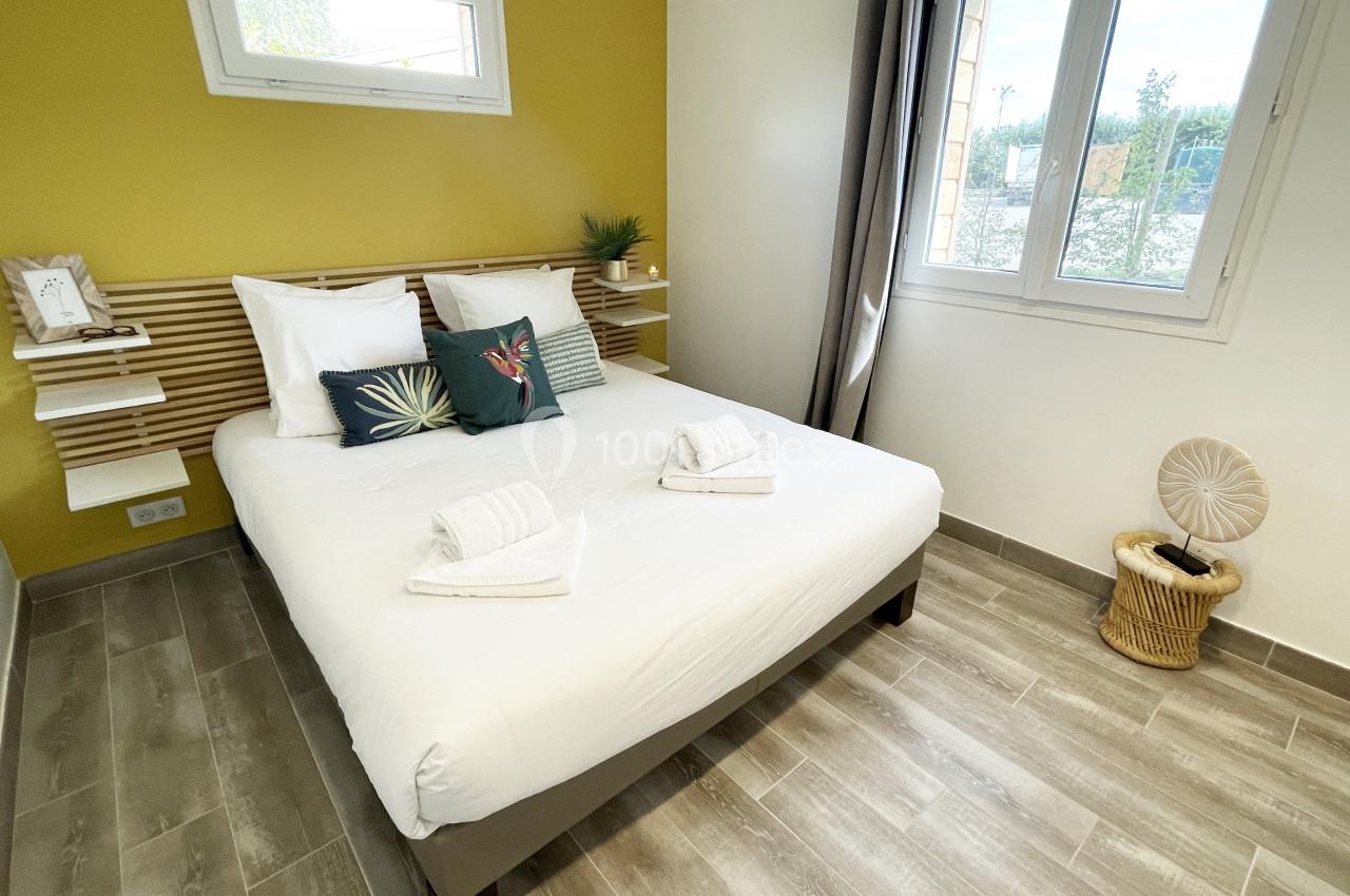 Chambre lumineuse avec un lit double, draps blancs, mur jaune, fenêtres et sol en carrelage gris.