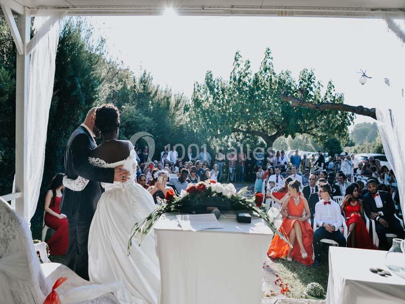 Un couple en tenue de mariage fait face à des invités assis lors d'une cérémonie en plein air, sous un ciel ensoleillé.