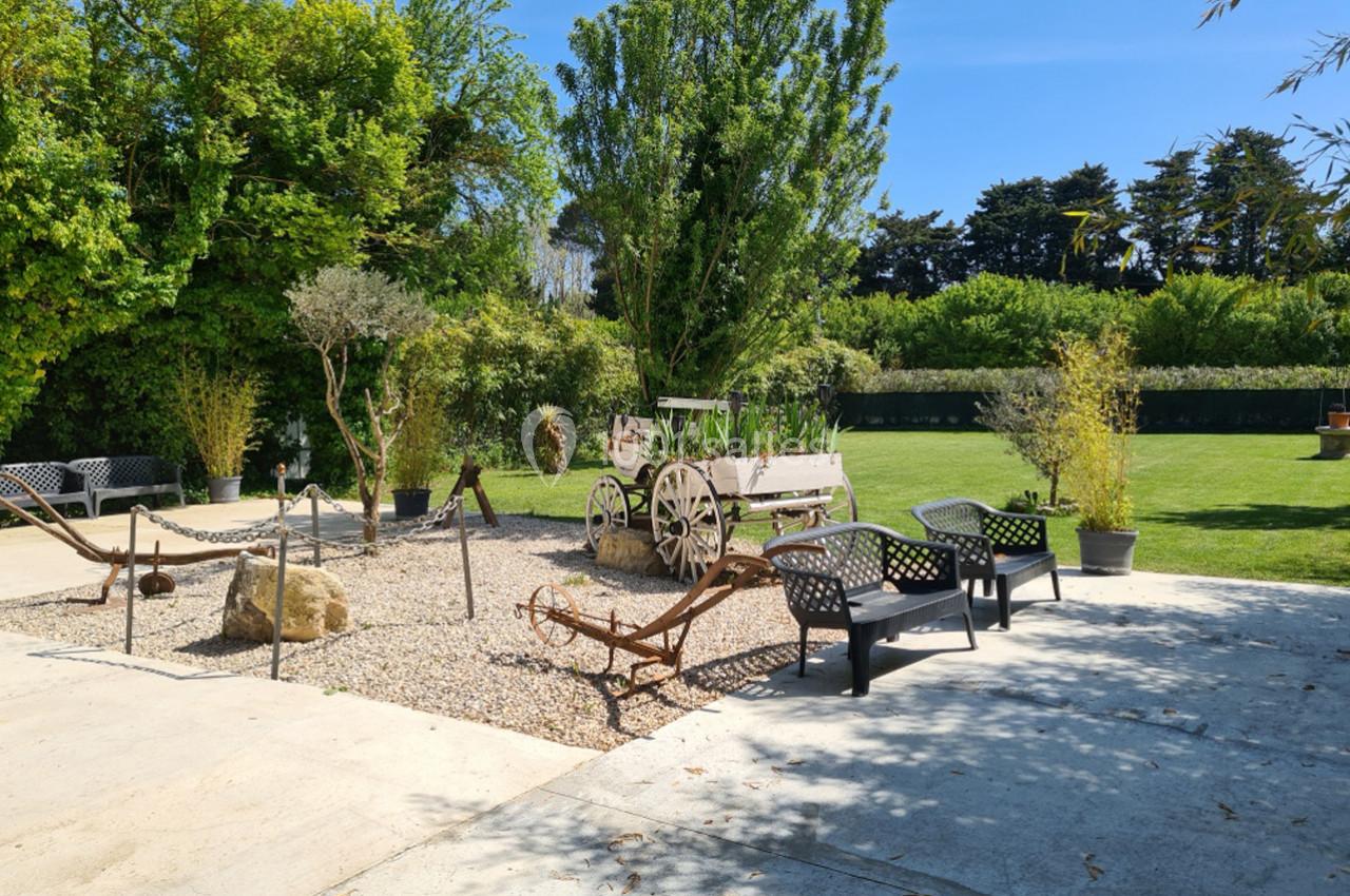 Espace extérieur avec bancs, gravier, charrette ancienne et végétation sous un ciel bleu.