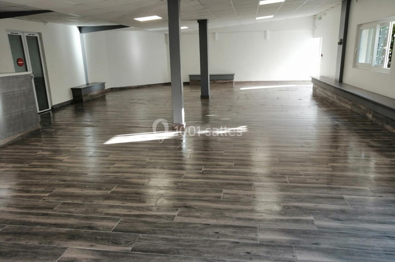 Salle vide avec un sol en parquet sombre, murs blancs, fenêtres et piliers gris sous un plafond à dalles blanches.