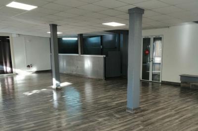 Salle vide avec un sol en parquet sombre, murs blancs, fenêtres et piliers gris sous un plafond à dalles blanches.