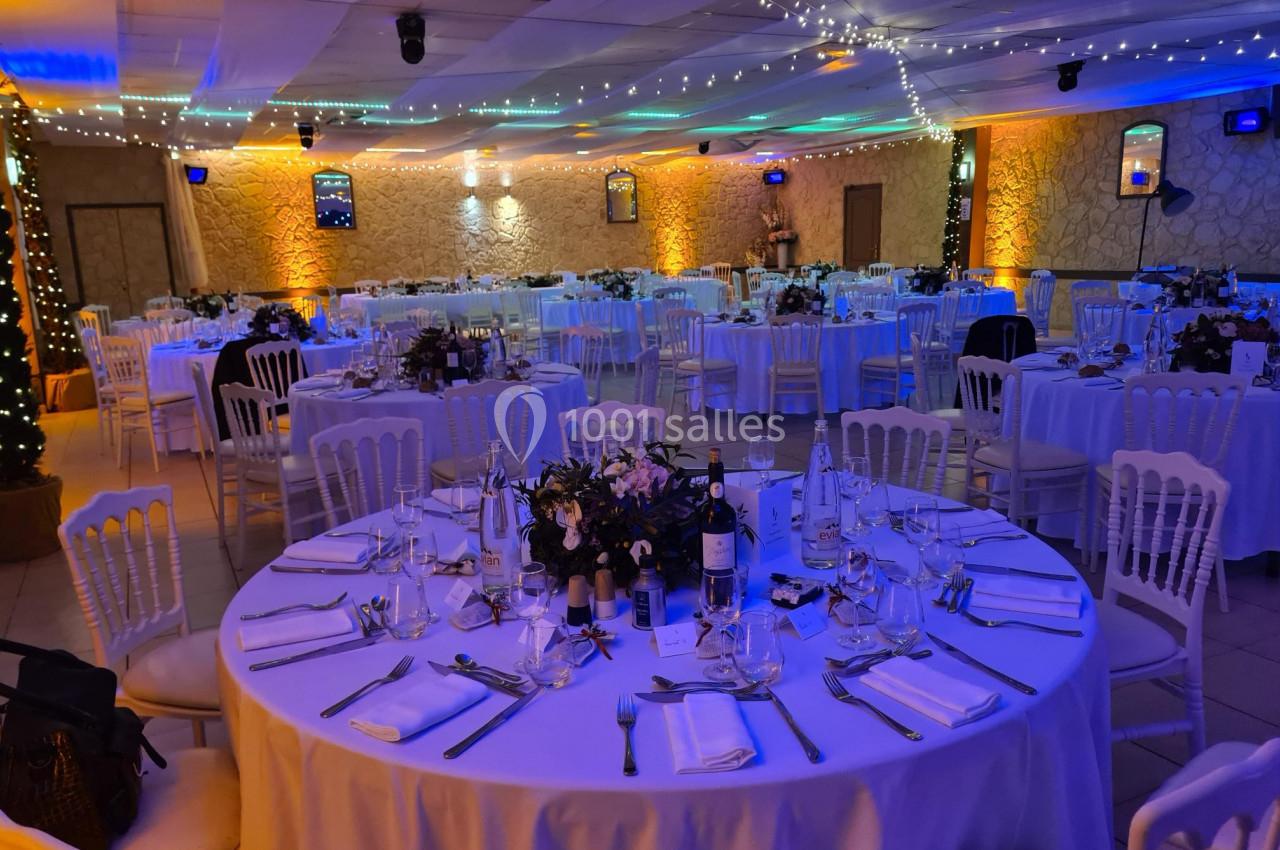 Salle de réception décorée avec des tables rondes dressées, éclairage tamisé et guirlandes lumineuses au plafond.