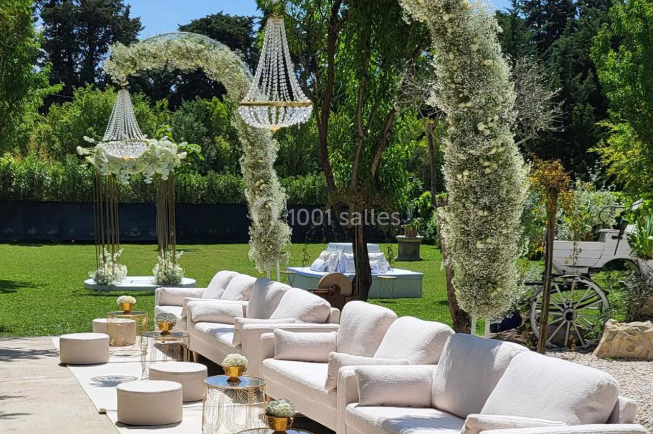 Espace extérieur décoré pour un événement avec canapés blancs, arches fleuries et lustres suspendus.
