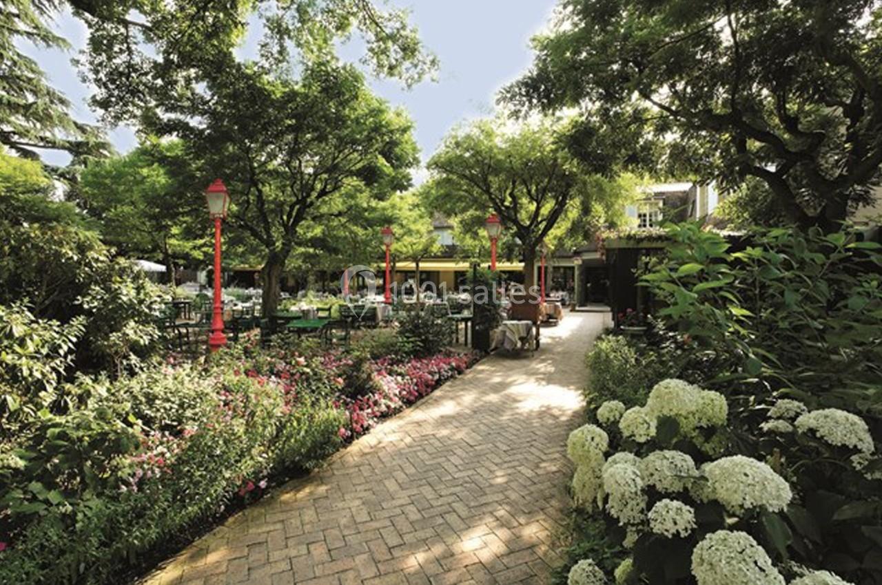 Allée pavée bordée de fleurs et d'arbustes, menant à une terrasse ombragée entourée d'arbres.