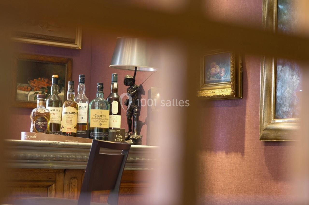 Bouteilles d'alcool alignées sur un comptoir en bois, entourées de cadres et d'une lampe dans un décor chaleureux.