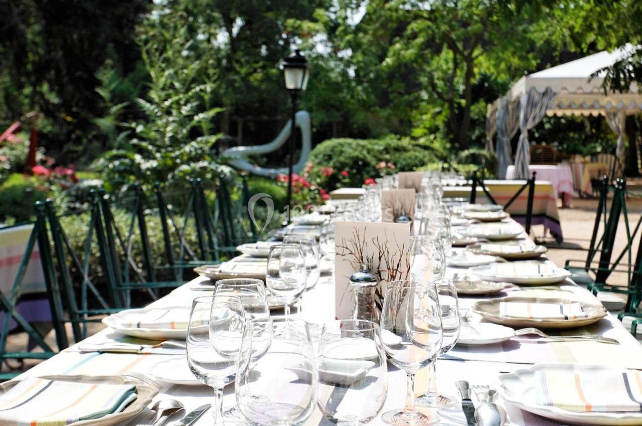 Table dressée en extérieur avec nappes blanches, verres et couverts, entourée de chaises dans un jardin verdoyant.
