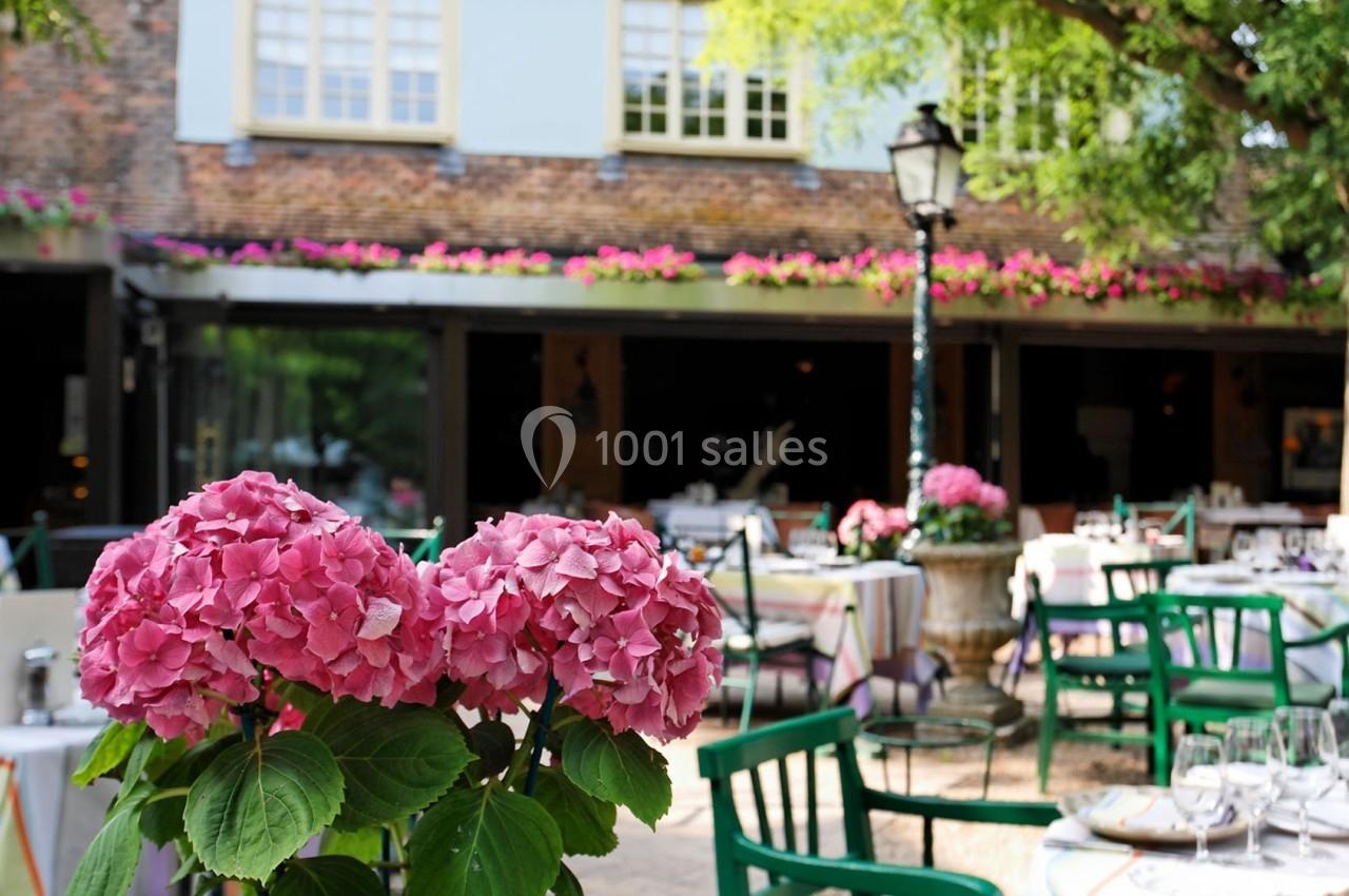Terrasse d'un restaurant avec des tables dressées, des chaises vertes et des hortensias roses en premier plan.