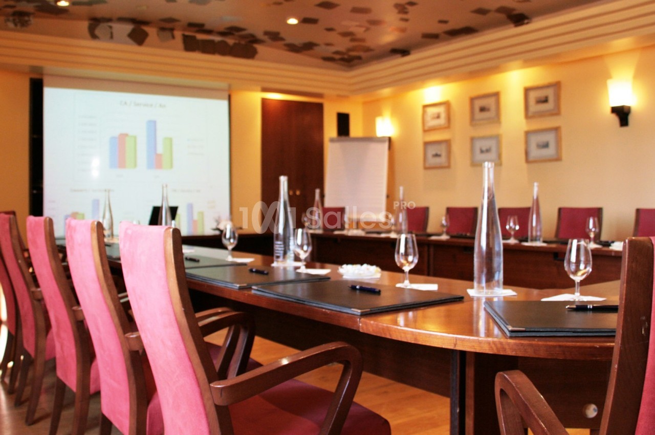 Salle de réunion avec une grande table en bois, chaises rouges, verres, blocs-notes et écran de présentation allumé.