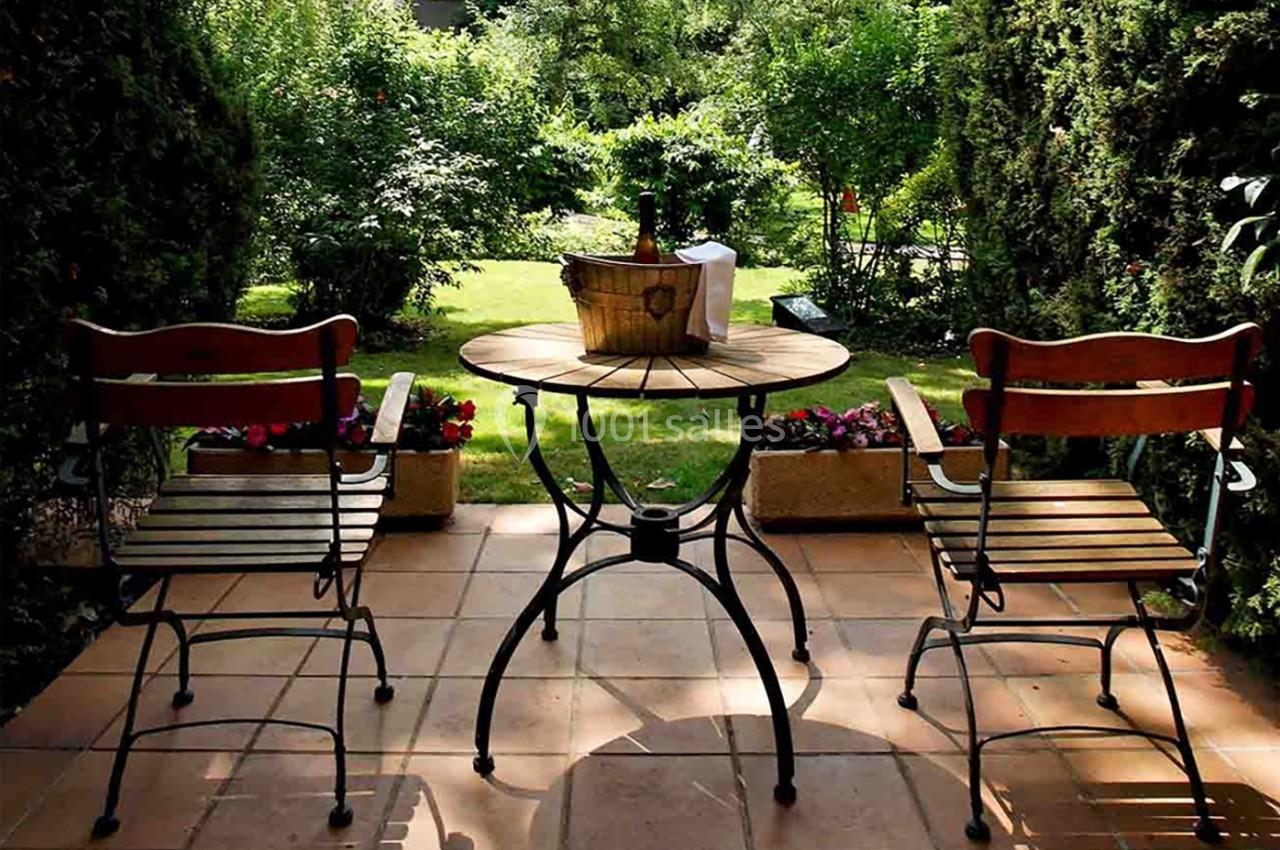 Deux chaises en bois et une table ronde avec un seau à glace sur une terrasse donnant sur un jardin verdoyant.