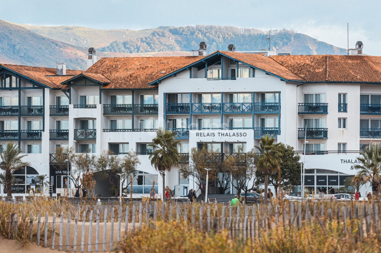 Façade d'un hôtel blanc avec balcons bleus, entouré de palmiers, situé près de dunes et de montagnes en arrière-plan.