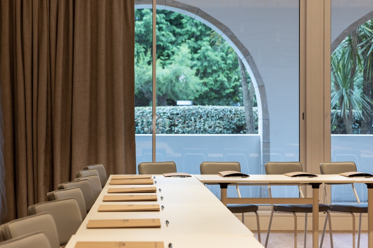 Salle de réunion lumineuse avec une grande table en U, des chaises grises et une vue sur un jardin à travers des baies…