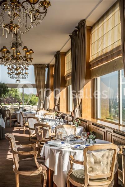 Salle de restaurant lumineuse avec grandes fenêtres, tables dressées et chaises en bois, décorée de lustres élégants.