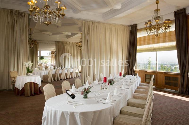 Salle de réception élégante avec tables dressées, nappes blanches, chandeliers et vue sur l'extérieur par une grande fenêtre.